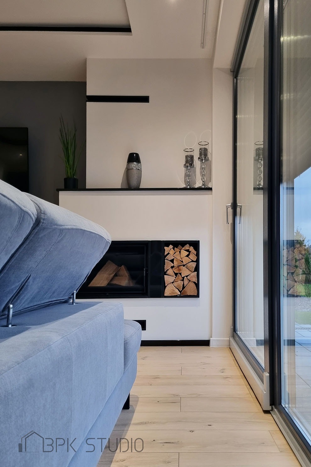 Jasne wnętrze salonu z kominkiem i drewnem, szary narożnik z podniesionym siedziskiem, duże okno przesuwne z widokiem na zewnątrz, dekoracje na półce nad kominkiem