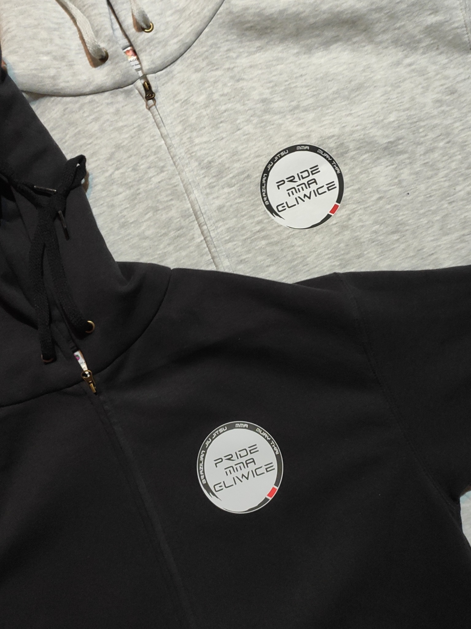 Czarne i szare bluzy z kapturem, zapinane na zamek, z okrągłym logo 'Pride MMA Gliwice' na piersi.