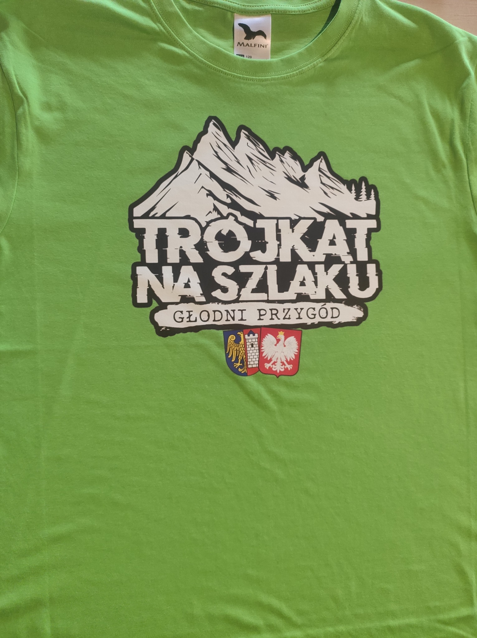Zielona koszulka z nadrukiem gór, napisu 'Trójkąt na szlaku' i herbów. Widoczna metka Malfini. Koszulka leży na płaskiej powierzchni.