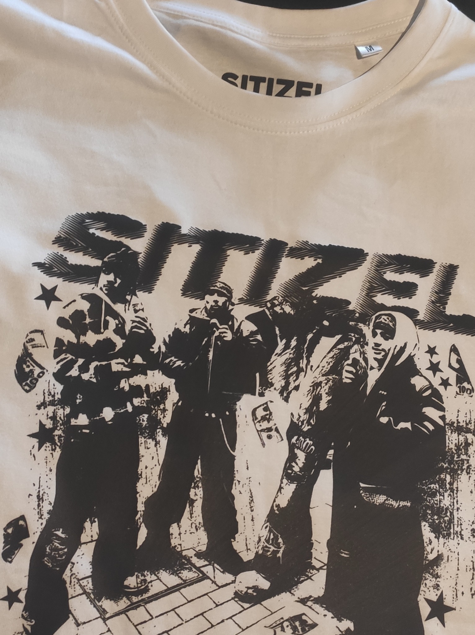 Biała koszulka z czarnym nadrukiem zespołu muzycznego z Gliwic. Grafika z wizerunkami muzyków, gwiazdami i banknotami. Widoczny fragment kołnierzyka i metka rozmiaru M.