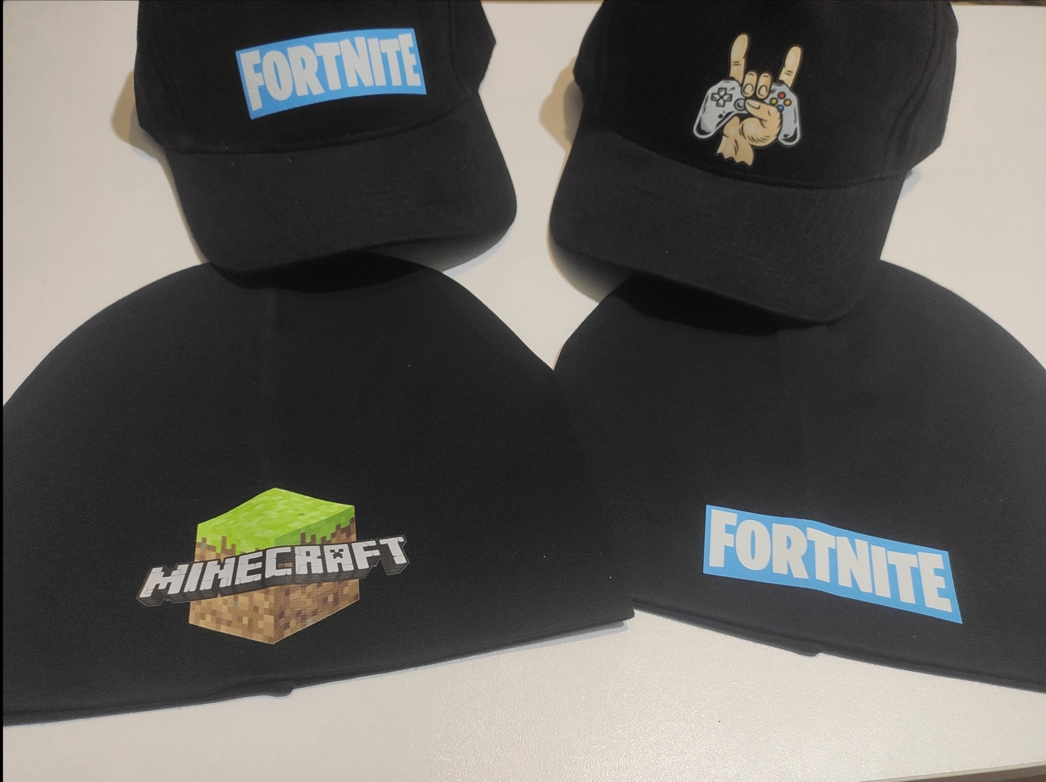 Czapki i czapki zimowe z nadrukami Minecraft i Fortnite, leżące na białym tle. Dwie czapki z daszkiem i dwie czapki bez daszka. Nadruki w stylu gamingowym.