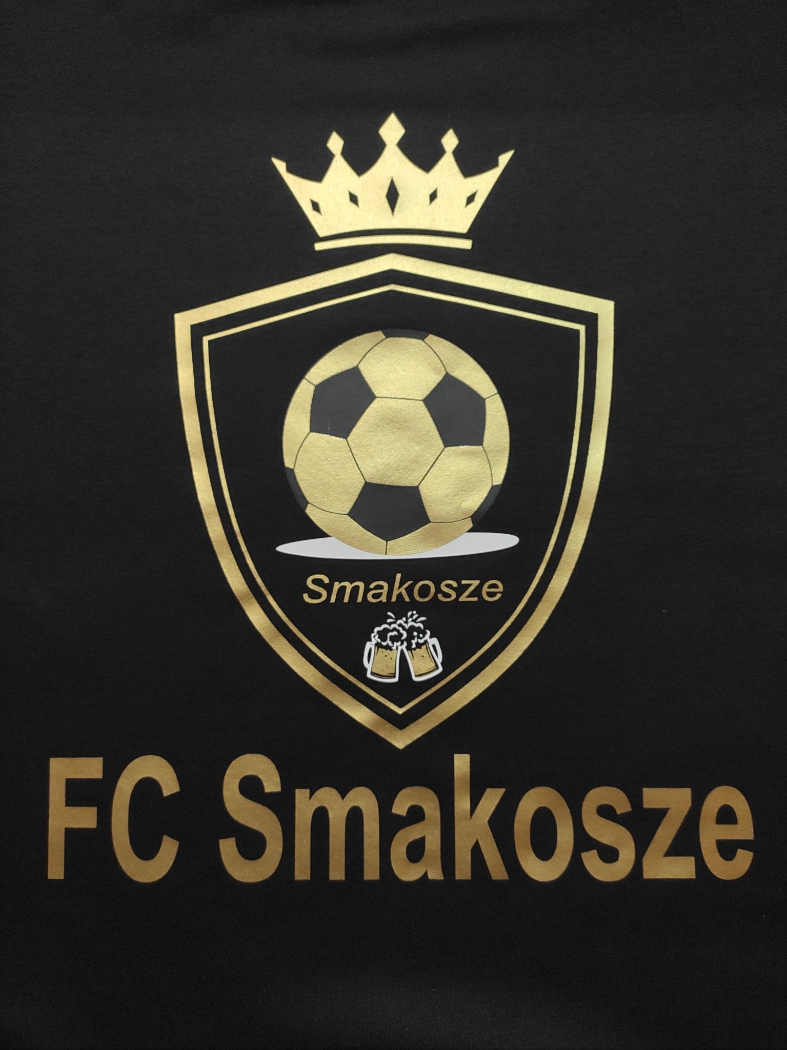 Czarna koszulka z złotym nadrukiem herbu drużyny piłkarskiej 'FC Smakosze' z piłką, koroną i kufelkami piwa.