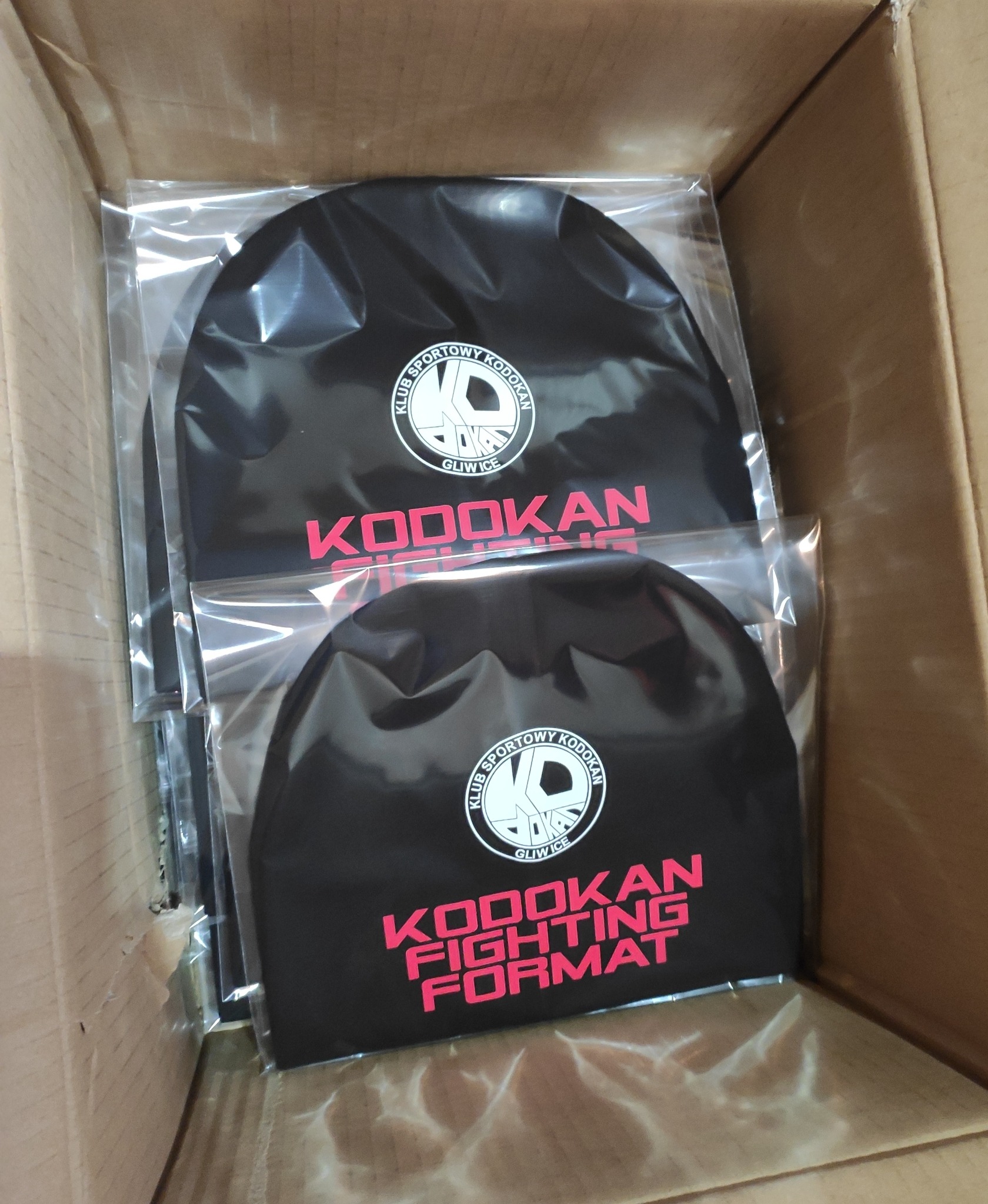 Czarne czapki z nadrukiem logo klubu sportowego Kodokan Fighting Format z Gliwic, zapakowane w foliowe woreczki i umieszczone w kartonowym pudełku.