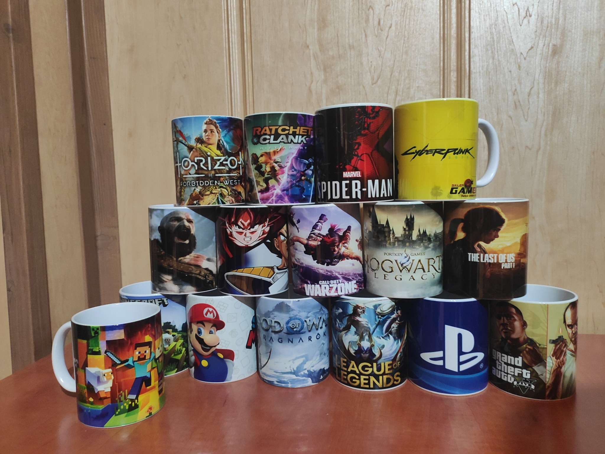 Kolekcja kubków z nadrukami motywów z gier komputerowych, ułożonych warstwowo na drewnianym stole, z widocznymi tytułami gier: Horizon Forbidden West, Ratchet & Clank, Marvel Spider-Man, Cyberpunk...