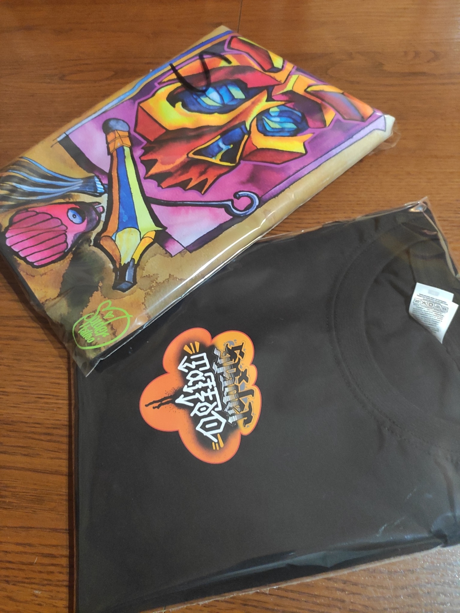 Dwie zapakowane koszulki z nadrukami leżą na drewnianym stole. Na jednej koszulce widoczny kolorowy, fantazyjny wzór, na drugiej czarnej koszulce pomarańczowy, stylizowany napis.