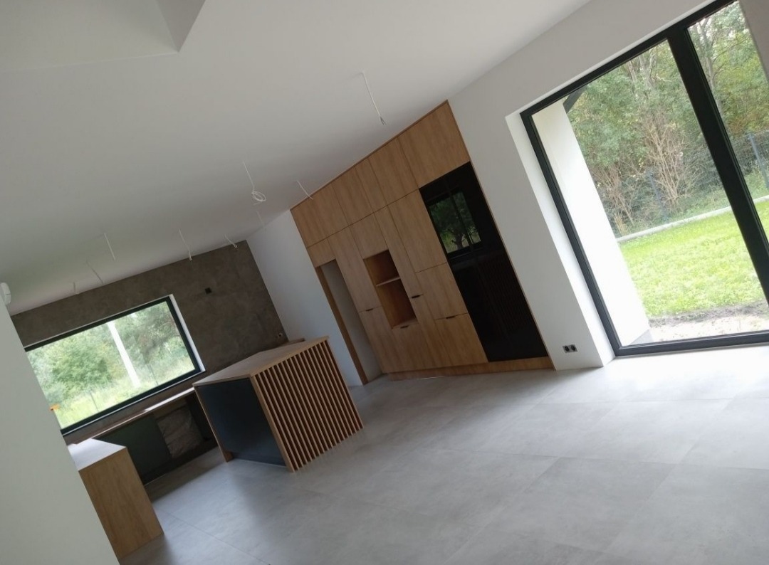 Jasne wnętrze z drewnianą zabudową kuchenną, wyspą z lamelami i dużym oknem z widokiem na zieleń, szara podłoga z płytek