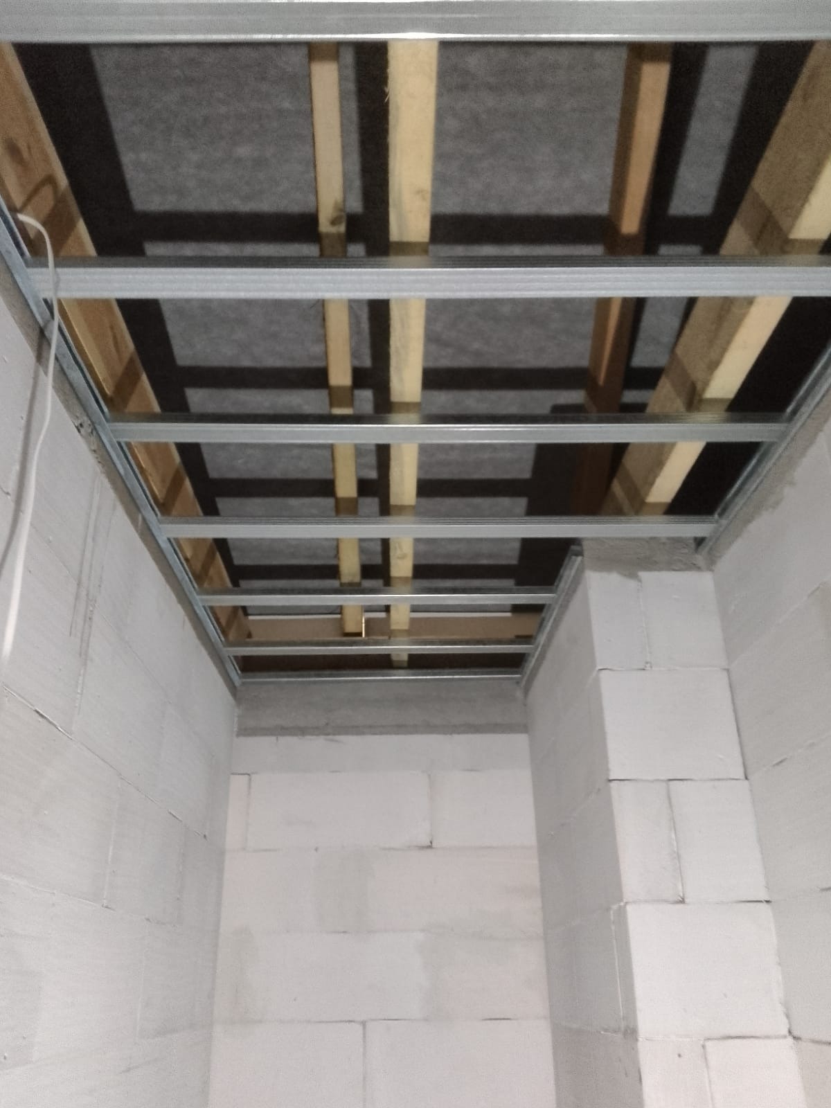 Stelaż sufitu podwieszanego z profili metalowych i drewnianych łat, widoczna izolacja z wełny mineralnej, ściany z białego betonu komórkowego.