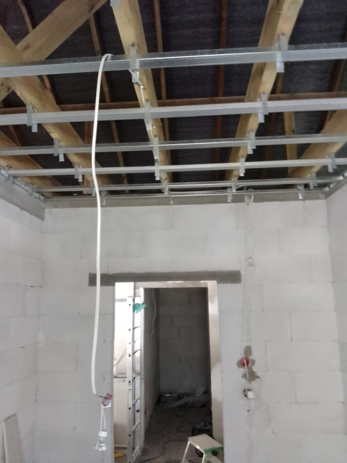 Konstrukcja metalowa podwieszanego sufitu zamontowana do drewnianych belek stropowych w surowym pomieszczeniu z białych bloczków, widoczne przewody elektryczne i drabina w przejściu.