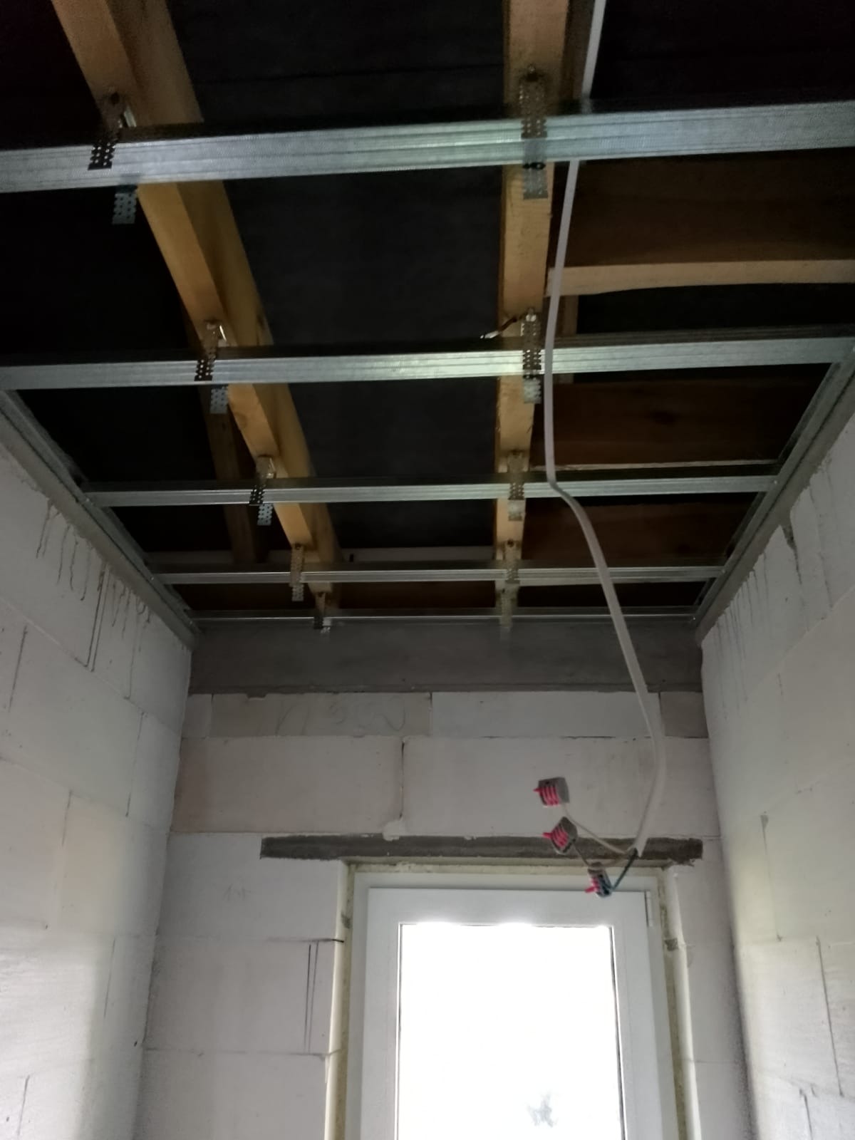 Montaż metalowej konstrukcji sufitu podwieszanego z widocznymi drewnianymi elementami nośnymi i instalacją elektryczną w surowym wnętrzu z białych bloczków.