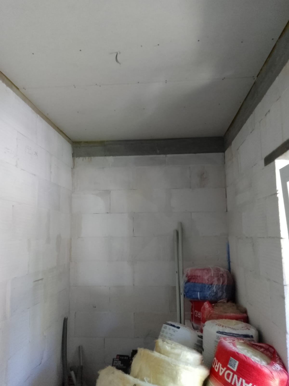 Wnętrze w trakcie prac wykończeniowych: ściany z białych bloczków, sufit z widocznymi punktami mocowania, worki z materiałami budowlanymi i wełna mineralna w rogu pomieszczenia.