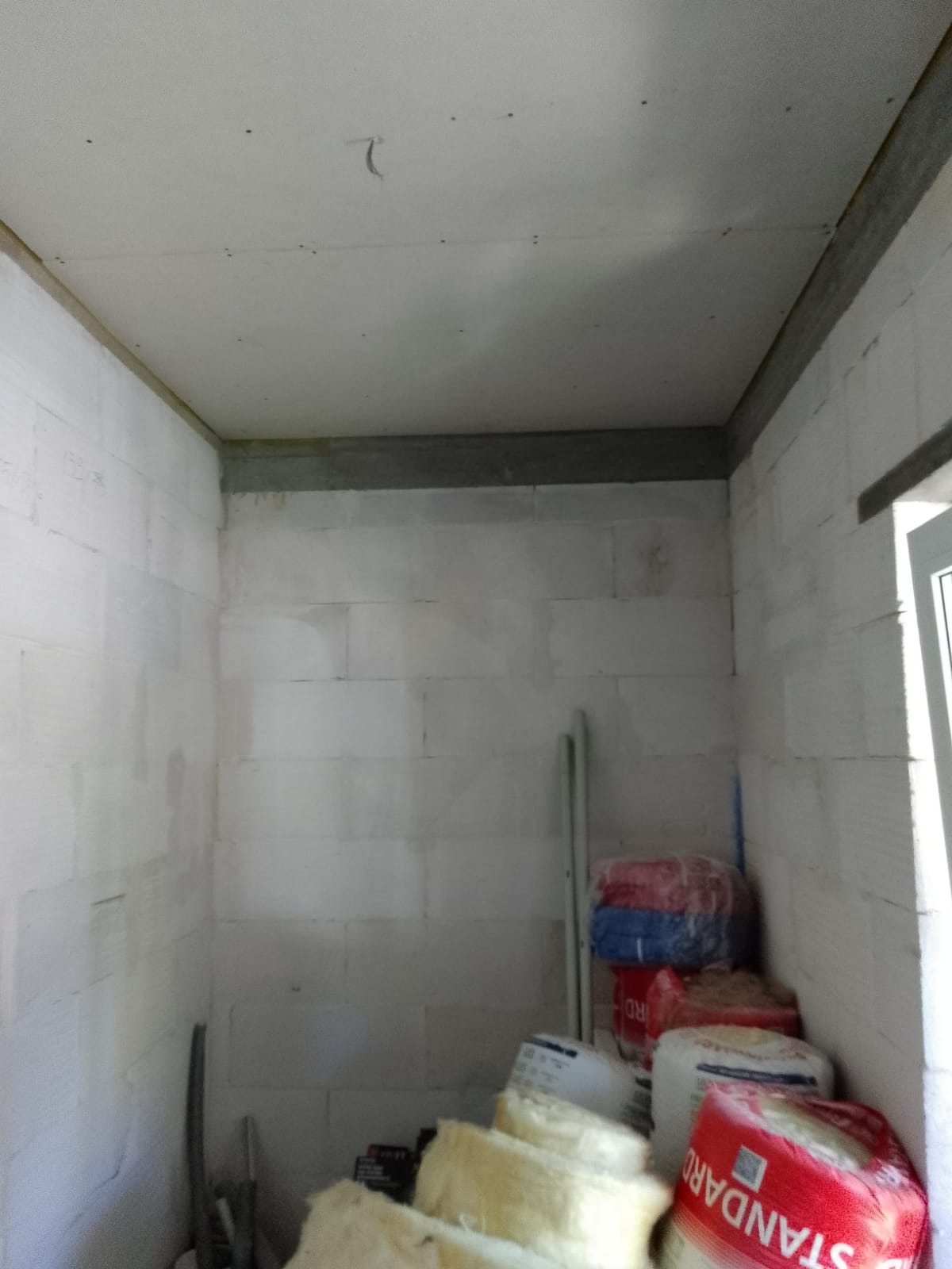 Wnętrze w trakcie prac wykończeniowych, widoczne ściany z bloków, ocieplony sufit z widocznymi przewodami oraz materiały budowlane w workach i rolkach.