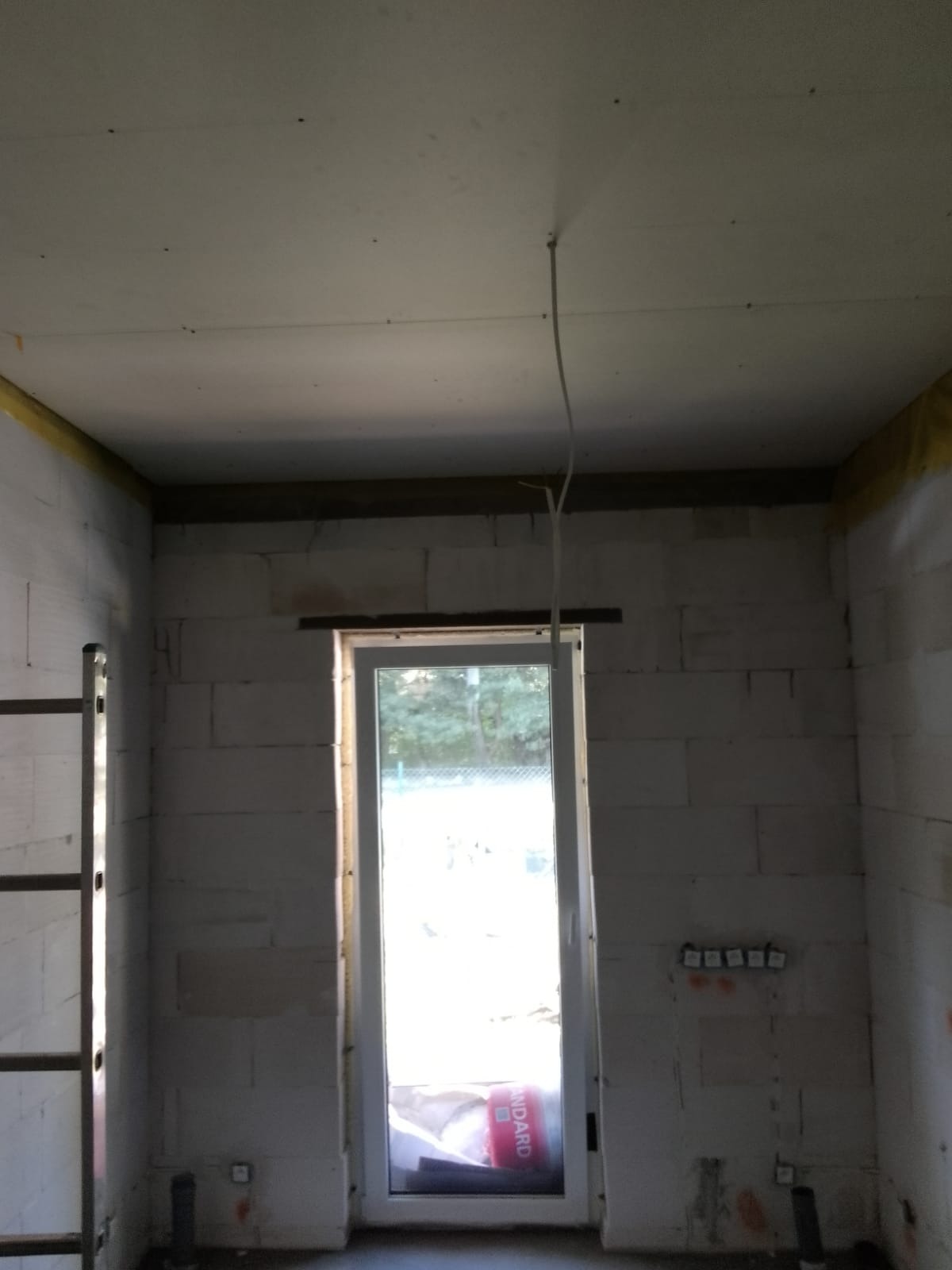 Surowy pokój w trakcie prac wykończeniowych: ściany z betonu komórkowego, okno z widokiem na zieleń, prowizorycznie zamontowany kabel na suficie, żółta taśma na łączeniu ściany z sufitem, drabina...