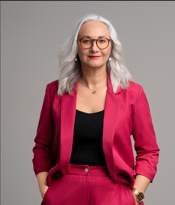 Portret kobiety w okularach i różowym garniturze, z rękami w kieszeniach, na szarym tle. Profesjonalny wizerunek biznesowy, z biżuterią i zegarkiem.