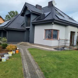 Dwukondygnacyjny dom z ciemnoszarą i białą elewacją, okno zabezpieczone folią malarską, widoczne puszki z farbą i rusztowanie.