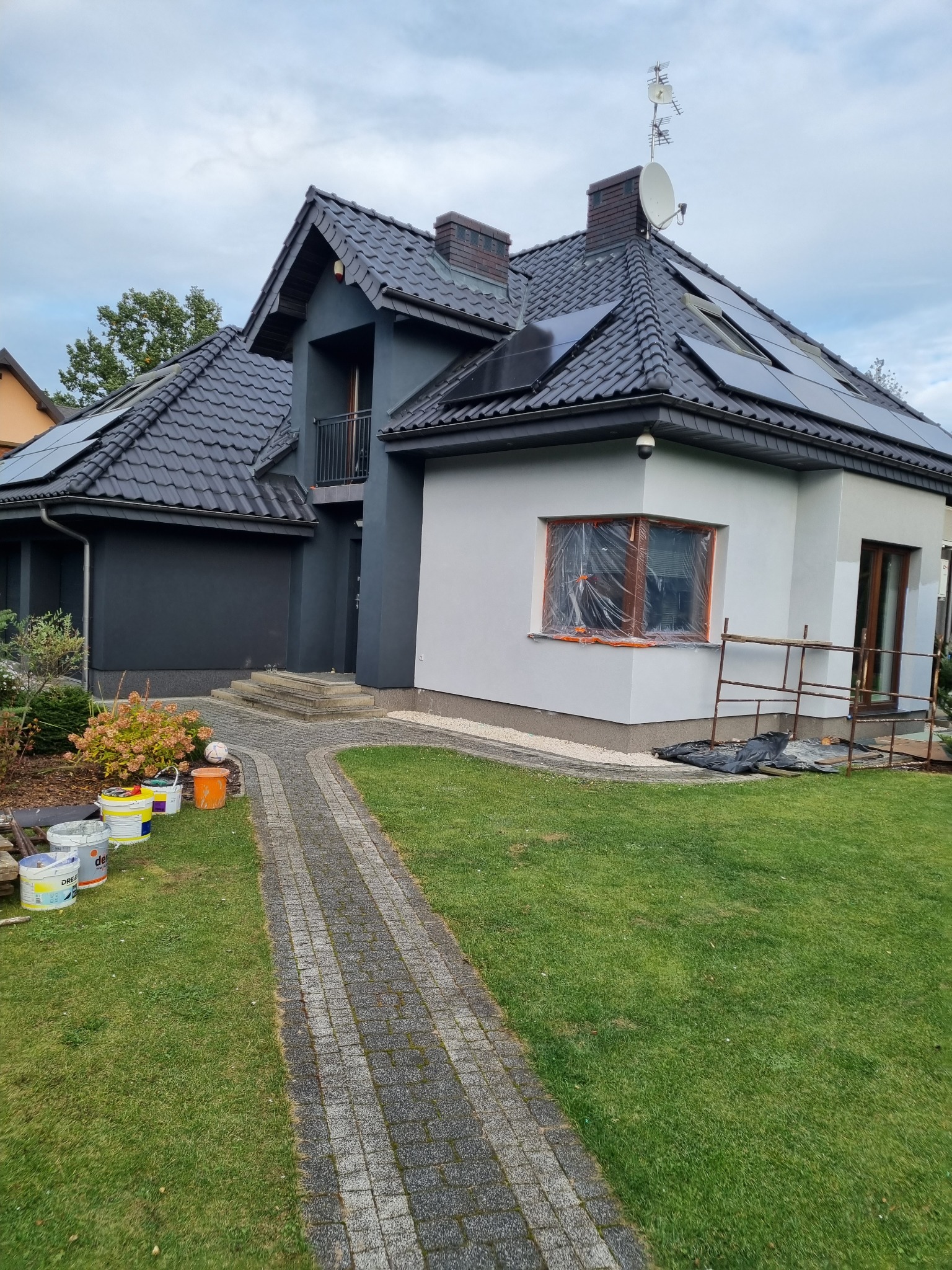 Dwukondygnacyjny dom z ciemnoszarą i białą elewacją, okno zabezpieczone folią malarską, widoczne puszki z farbą i rusztowanie.