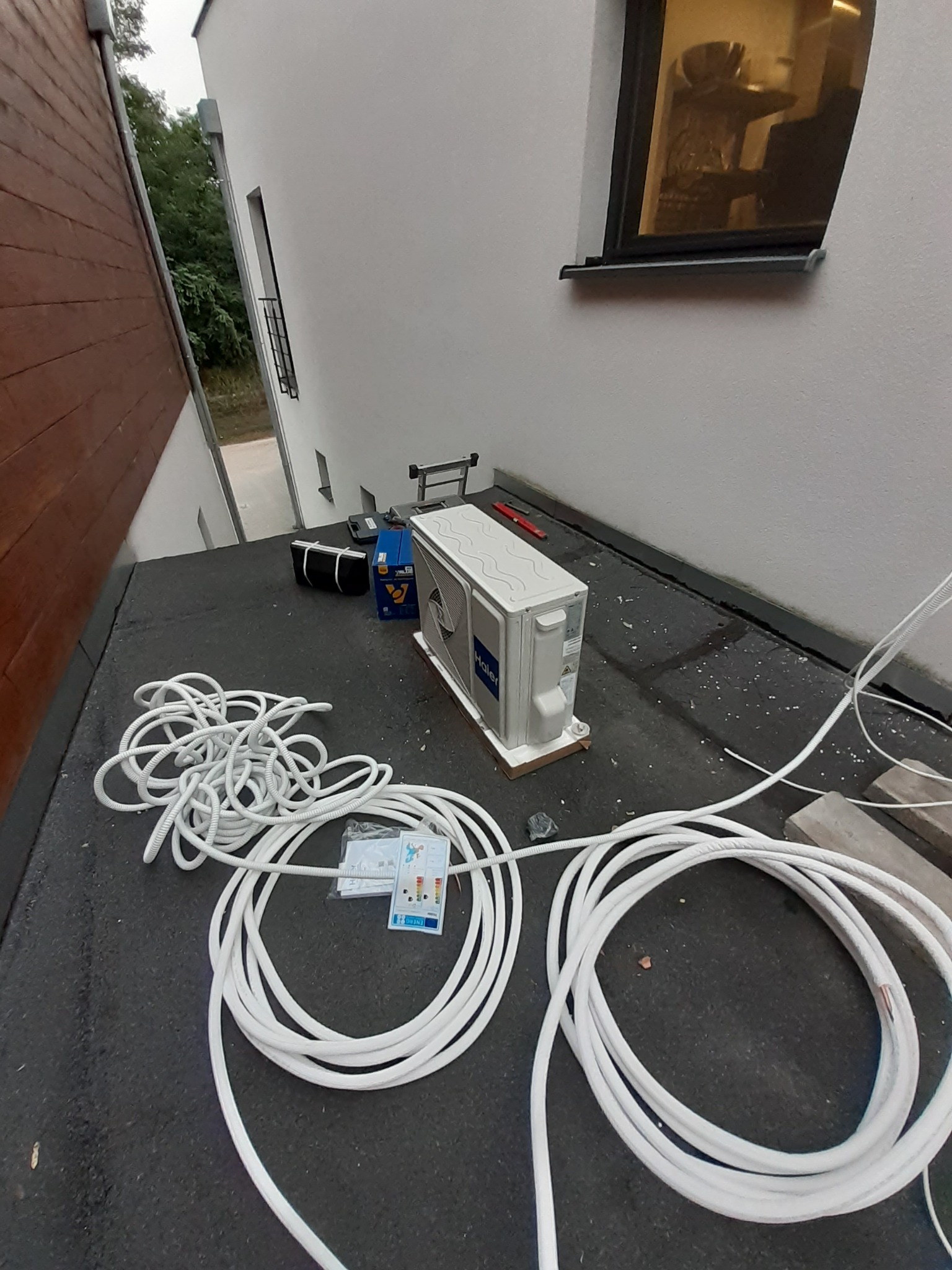 Przygotowanie do montażu klimatyzatora Haier na dachu budynku, widoczne zwinięte kable, skrzynka narzędziowa i instrukcja obsługi.
