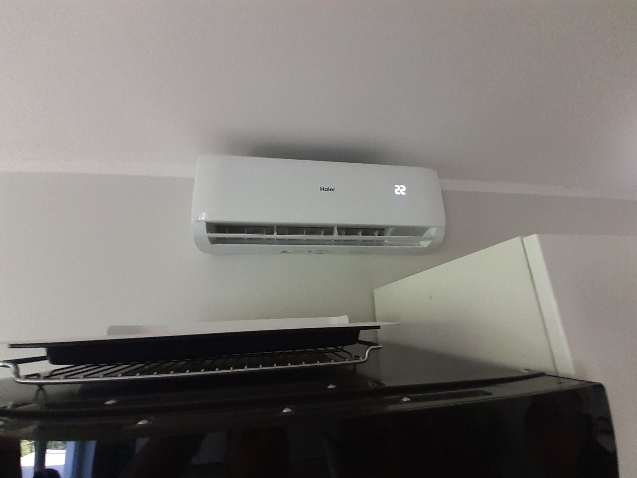 Biały klimatyzator Haier z wyświetlaczem temperatury 22 stopnie Celsjusza zamontowany na ścianie w pomieszczeniu. Pod spodem widoczny czarny blat z kratką i blachą.