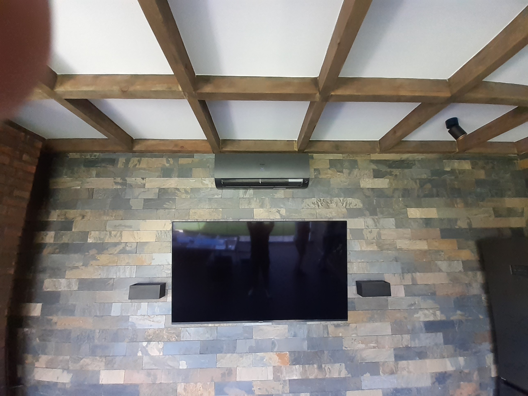 Szara jednostka klimatyzacyjna zamontowana nad telewizorem na ścianie z kamienia dekoracyjnego pod drewnianym sufitem kasetonowym.