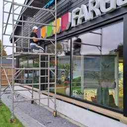 RENOWA BYGG MAGIERA - Pracownik na rusztowaniu montuje lub czyści wielokolorowy szyld z napisem 'FARGE'