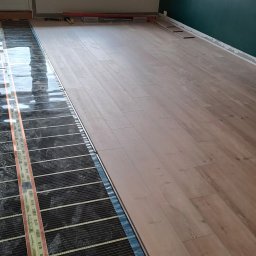 RENOWA BYGG MAGIERA - Montaż ogrzewania podłogowego: widoczna mata grzewcza z folią i częściowo ułożone panele podłogowe w jasnym odcieniu drewna, z oknem i balkonem w tle.