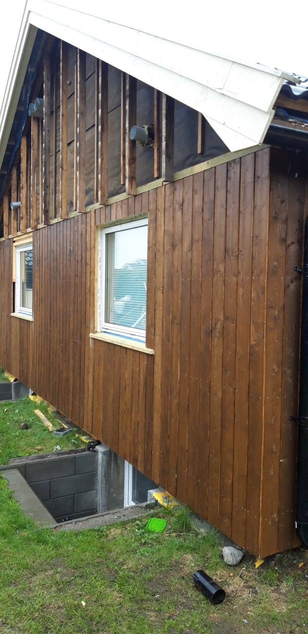 Dom w trakcie prac wykończeniowych elewacji zewnętrznej, widoczne drewniane deski pomalowane na brązowo, fragment ściany bez desek z odsłoniętą izolacją oraz dwa okna z białymi ramami.