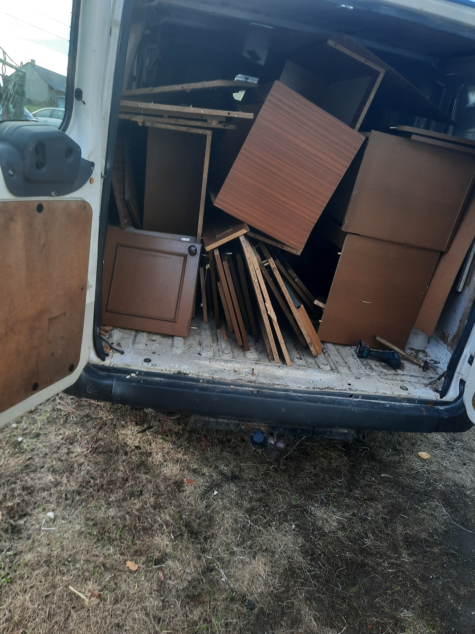 Wnętrze białego busa wypełnione elementami zdemontowanych mebli, w tym płyty drewniane i drzwiczki szafek w odcieniach brązu, widoczna wiertarka akumulatorowa i młotek, tło stanowi trawnik...