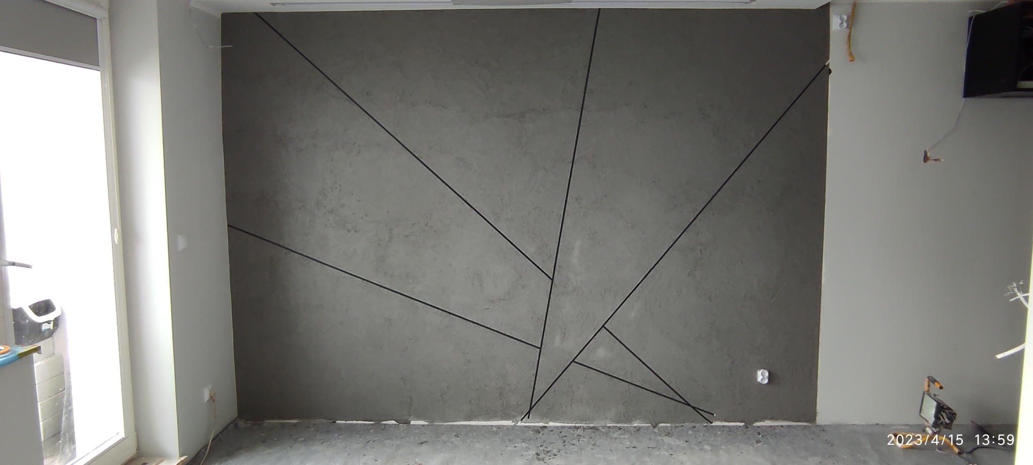 Ściana z szarą, dekoracyjną fakturą i czarnymi, geometrycznymi liniami, widoczne gniazdko elektryczne i fragment lampy budowlanej.