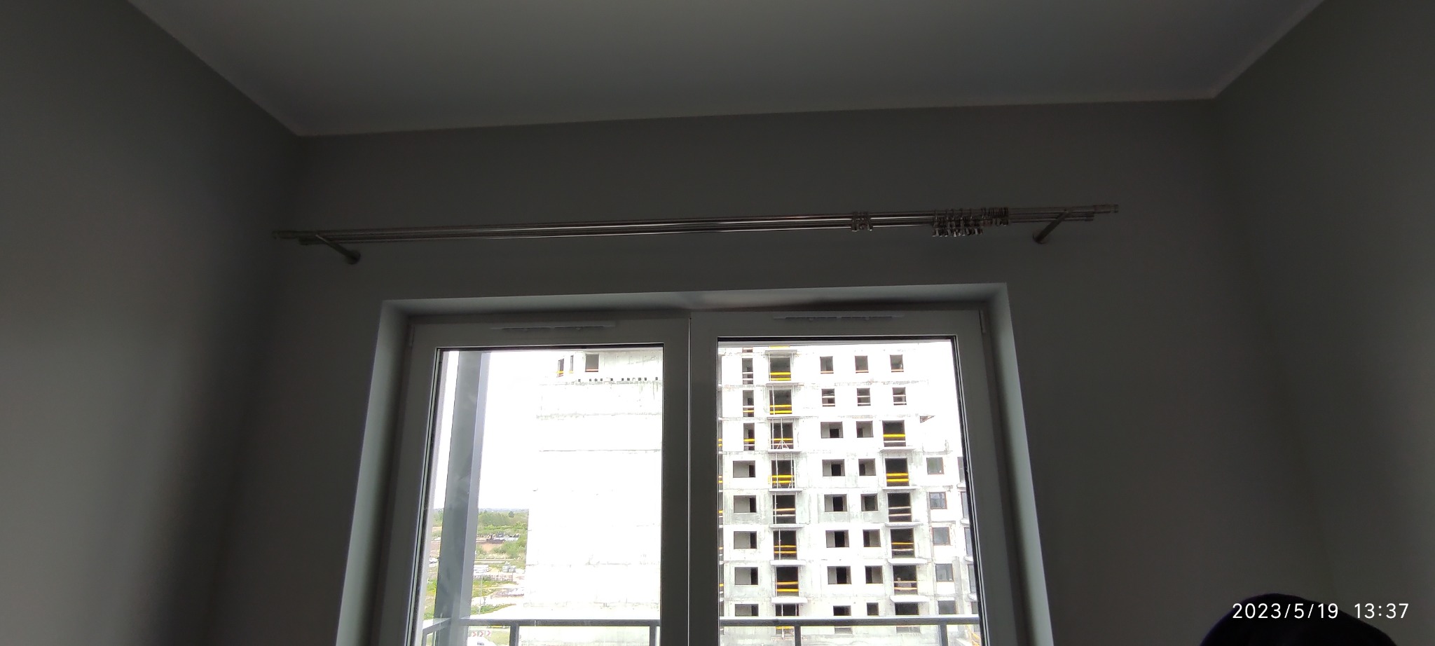 Zamontowany, podwójny karnisz metalowy z uchwytami i żabkami nad nowym, białym oknem z widokiem na budynek w stanie surowym.