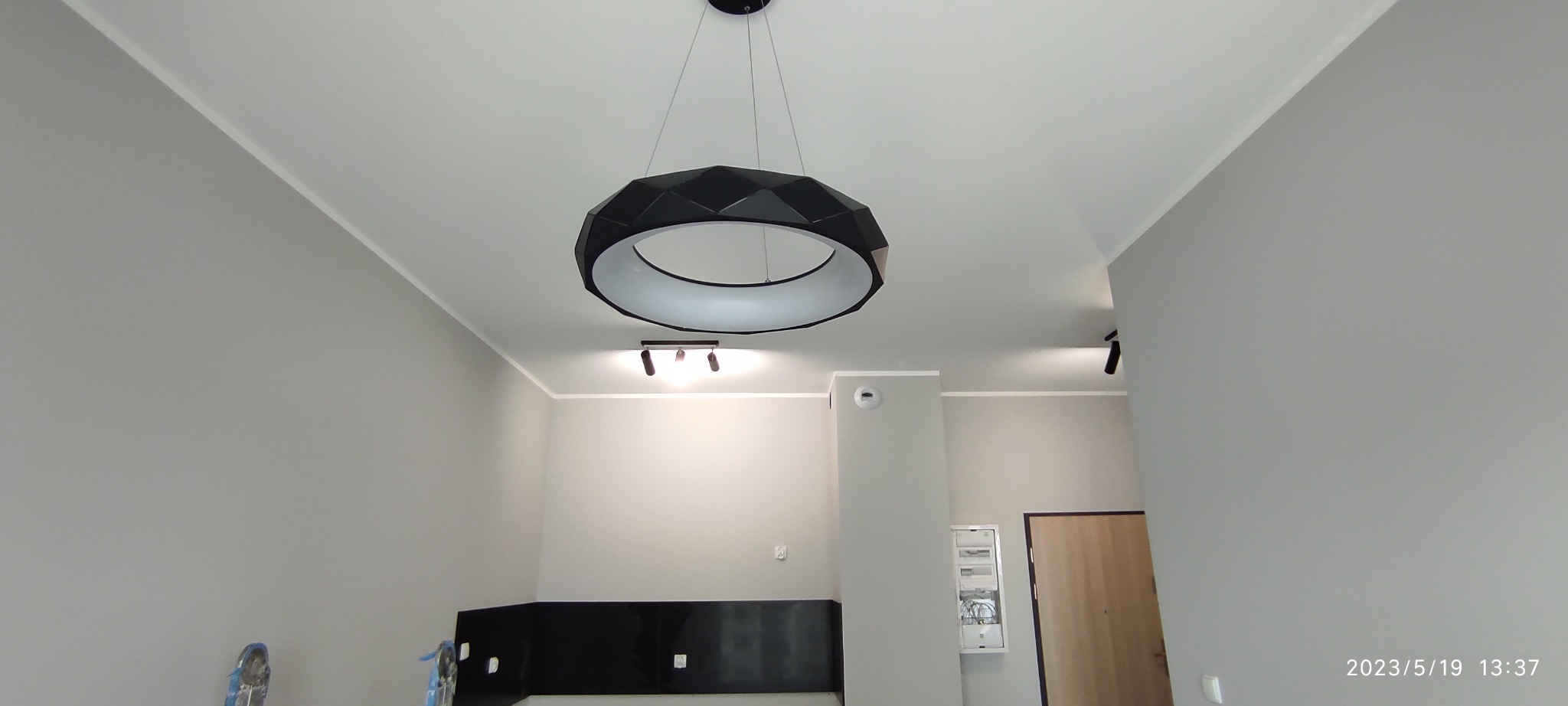 Nowoczesny, okrągły żyrandol z czarną, fasetowaną obudową i białym wnętrzem, zawieszony na cienkich linkach w szarym pomieszczeniu z listwami przysufitowymi i punktowym oświetleniem.