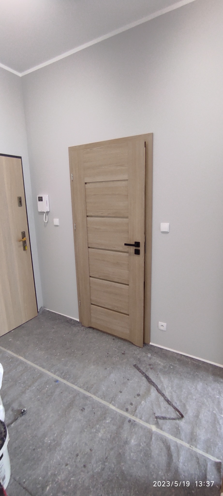 Jasne, drewnopodobne drzwi wewnętrzne z poziomymi panelami i czarną klamką, częściowo otwarte, widoczne na tle szarej ściany i podłogi w trakcie remontu, z widocznymi śladami po pracach.