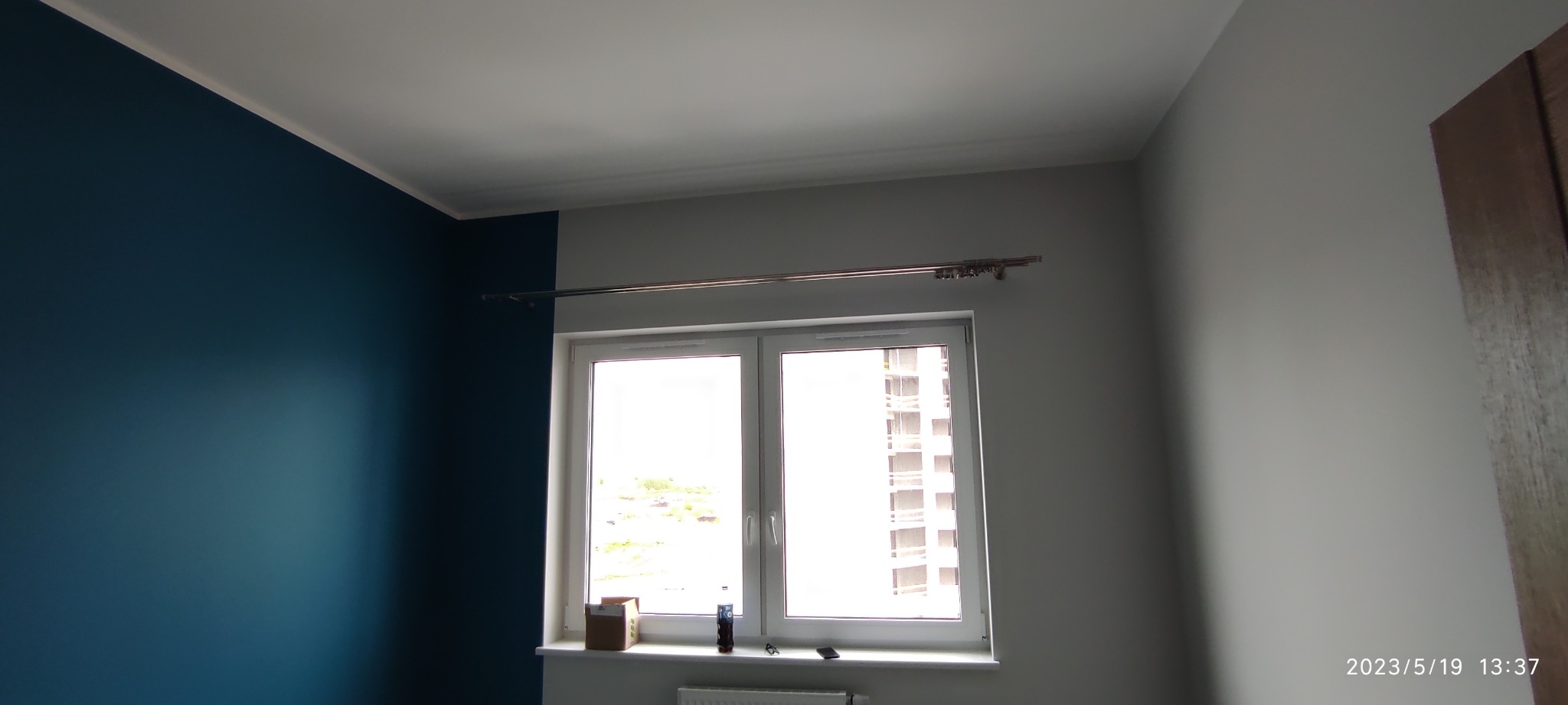 Pomalowane na granatowo i szaro ściany w pokoju z białym oknem i metalowym karniszem, widok na budynek zza okna.