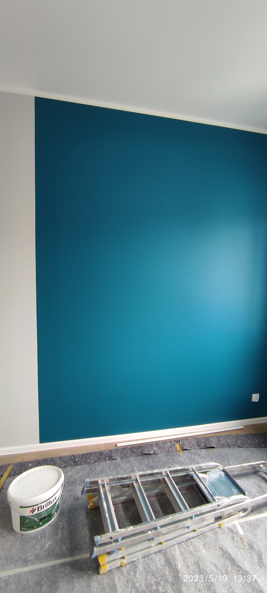 Świeżo pomalowana ściana na intensywny turkusowy kolor, z widocznym wiadrem z farbą marki Brillux i rozłożoną drabiną na podłodze zabezpieczonej folią malarską.