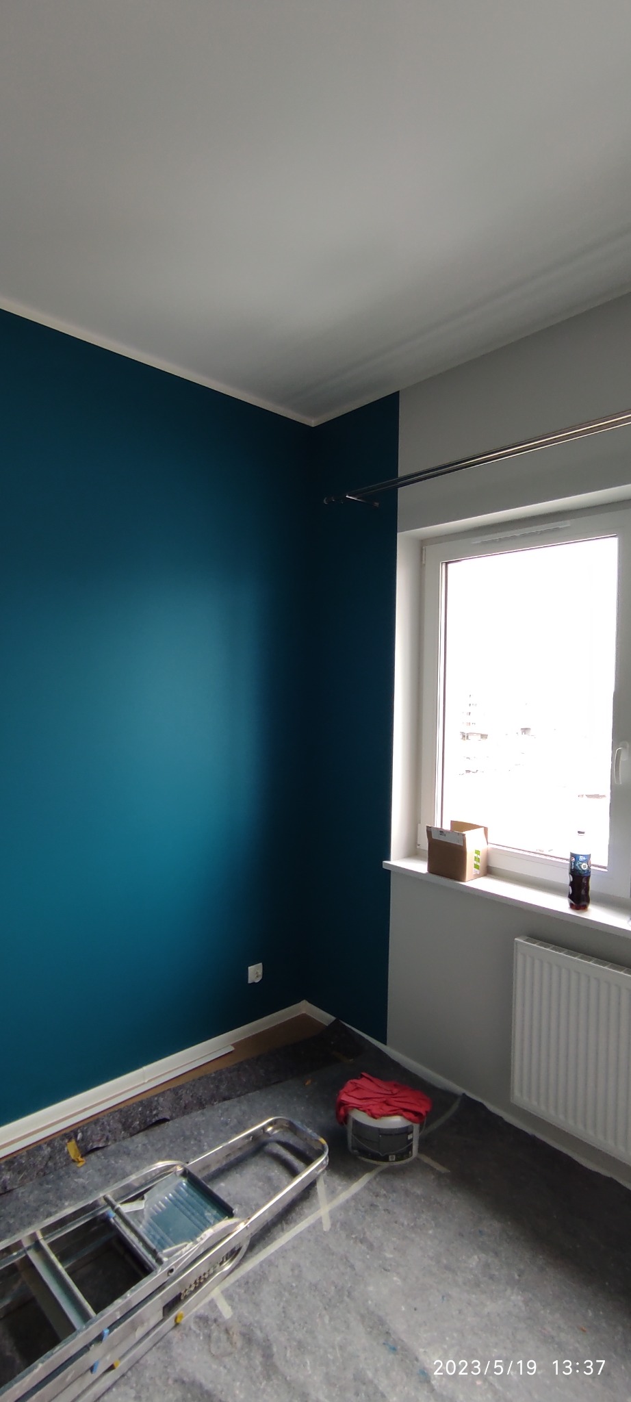 Pokój w trakcie malowania: ściana pomalowana na intensywny turkus, druga ściana szara, drabina malarska na podłodze zabezpieczonej folią, widoczne wiadro z farbą przykryte czerwoną szmatką.