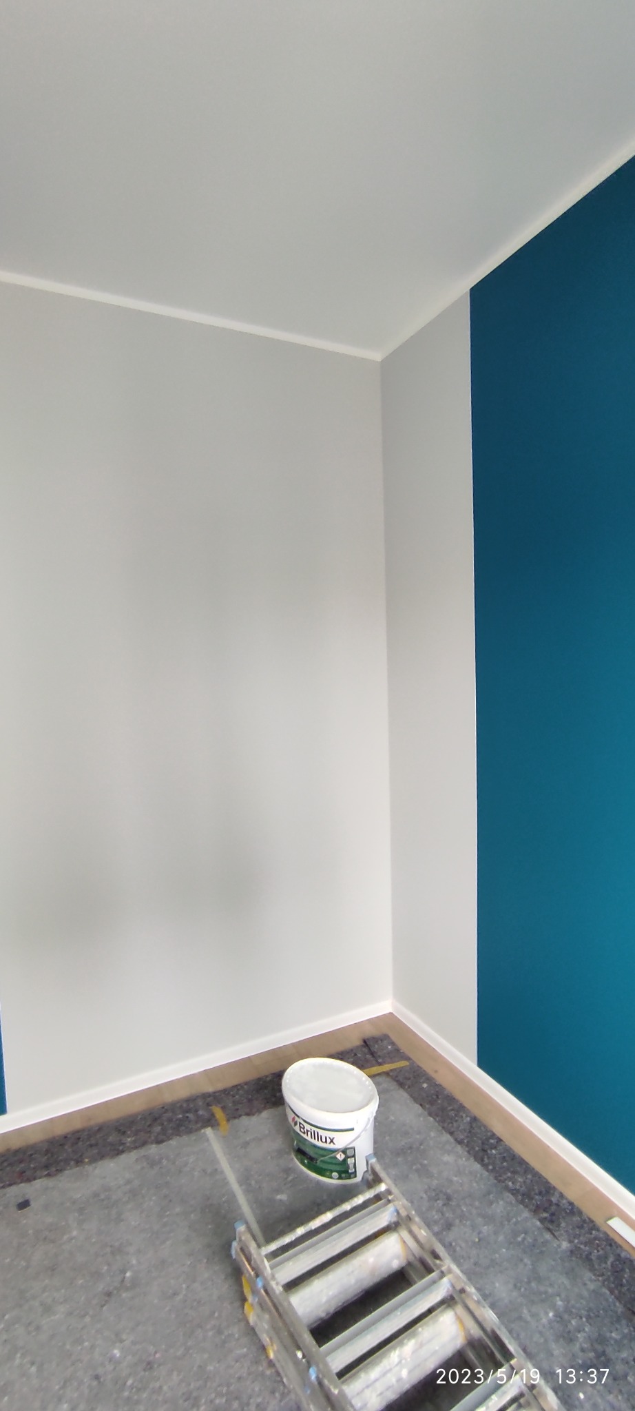 Świeżo pomalowany narożnik pokoju w odcieniach bieli i turkusu, widoczna drabina malarska i wiadro farby marki Brillux, podłoga zabezpieczona folią.