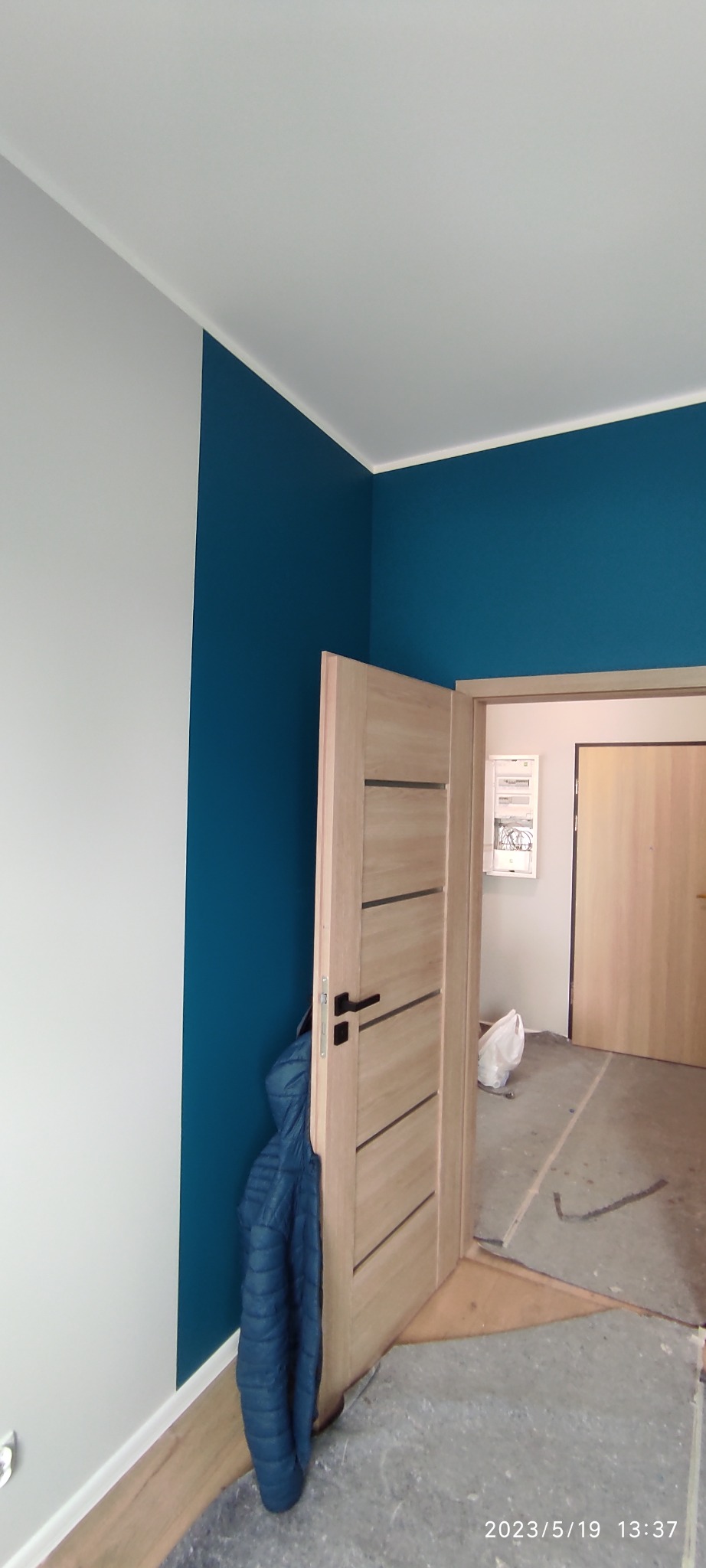 Świeżo zamontowane, jasne drzwi wewnętrzne z poziomymi frezowaniami, czarną klamką i niebieską kurtką wiszącą obok, na tle pomalowanych na biało i niebiesko ścian oraz podłogi w trakcie remontu.