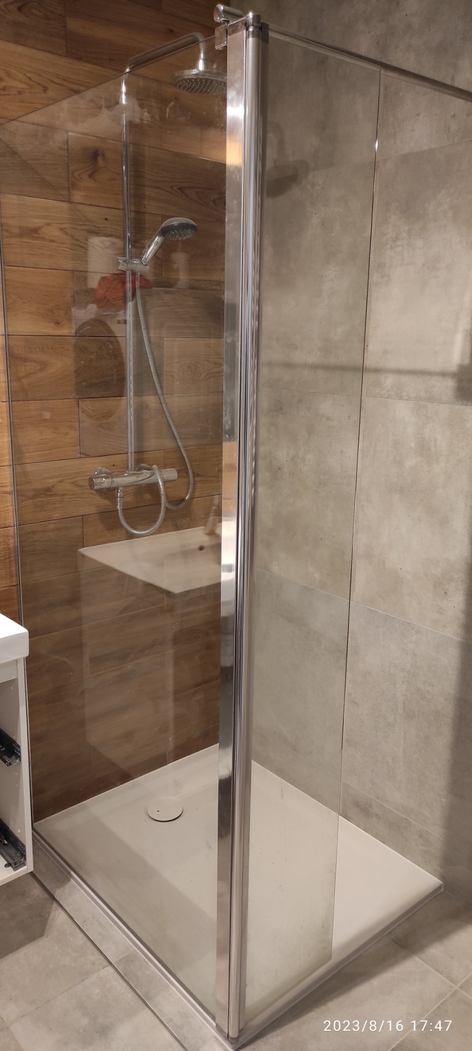 Kabina prysznicowa z chromowanymi profilami i szklanymi ścianami, widok na drewnopodobne płytki na ścianie i szarą płytkę na drugiej ścianie oraz biały brodzik.