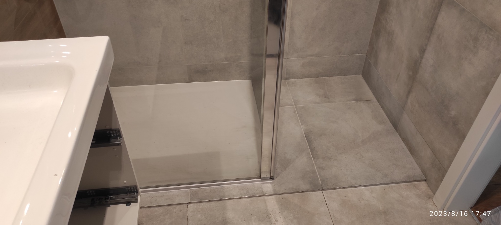 Fragment łazienki z widocznym brodzikiem z kabiną prysznicową, umywalką z szafką z wysuwanymi szufladami, oraz szarą płytką ceramiczną na ścianach i podłodze.