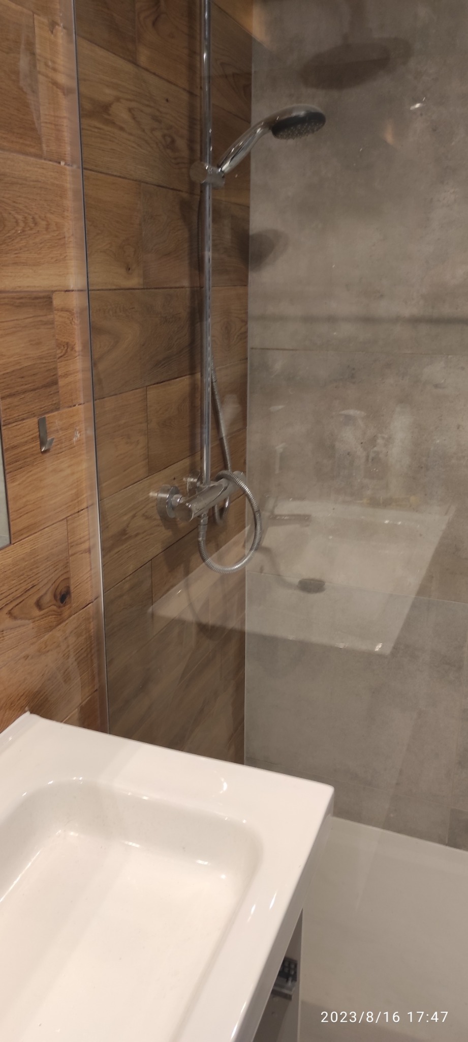 Nowoczesna kabina prysznicowa z deszczownicą i baterią termostatyczną, wykończona płytkami imitującymi drewno i beton, z widoczną białą umywalką w rogu kadru.