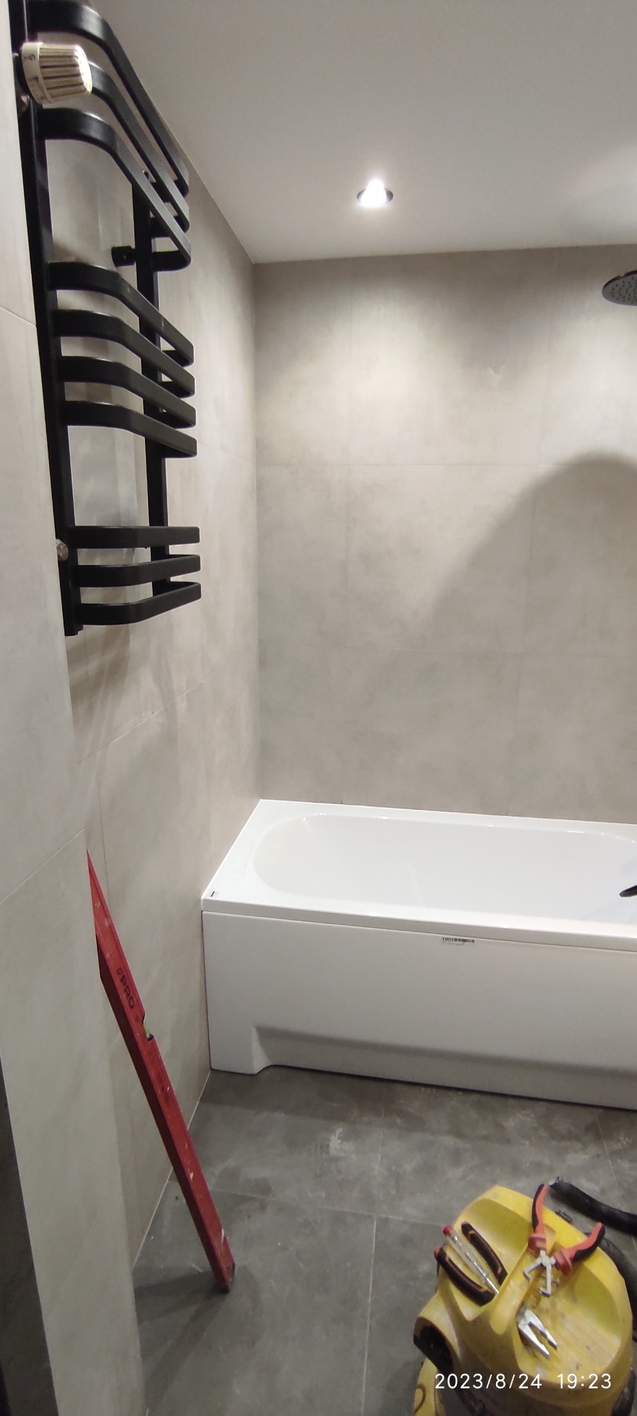 Łazienka w trakcie prac wykończeniowych: biała wanna, grzejnik drabinkowy w kolorze czarnym zamontowany na ścianie z szarymi płytkami, poziomica oparta o ścianę, na podłodze leżą narzędzia.