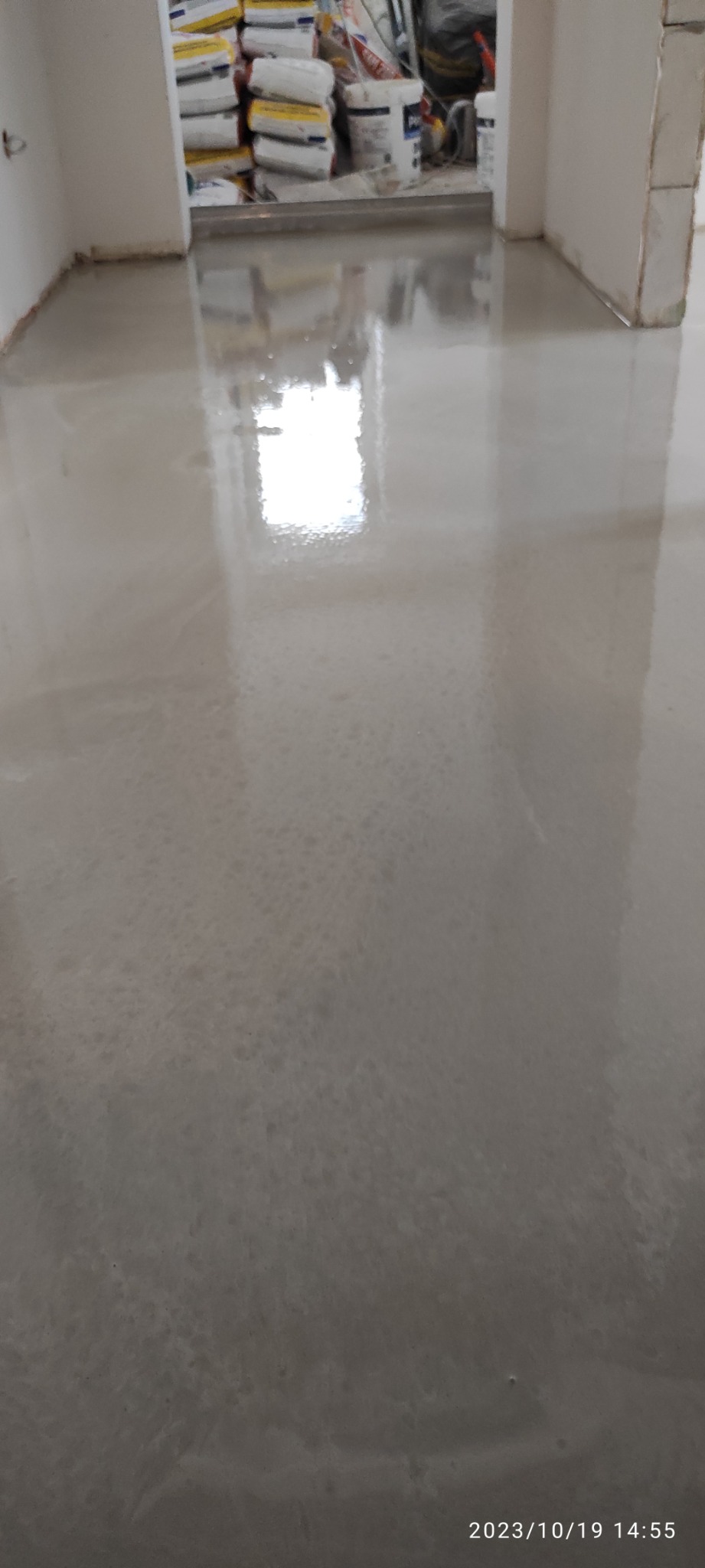 Świeżo wylana, samopoziomująca wylewka podłogowa, jeszcze mokra, odbijająca światło, z widocznymi workami z zaprawą w tle.