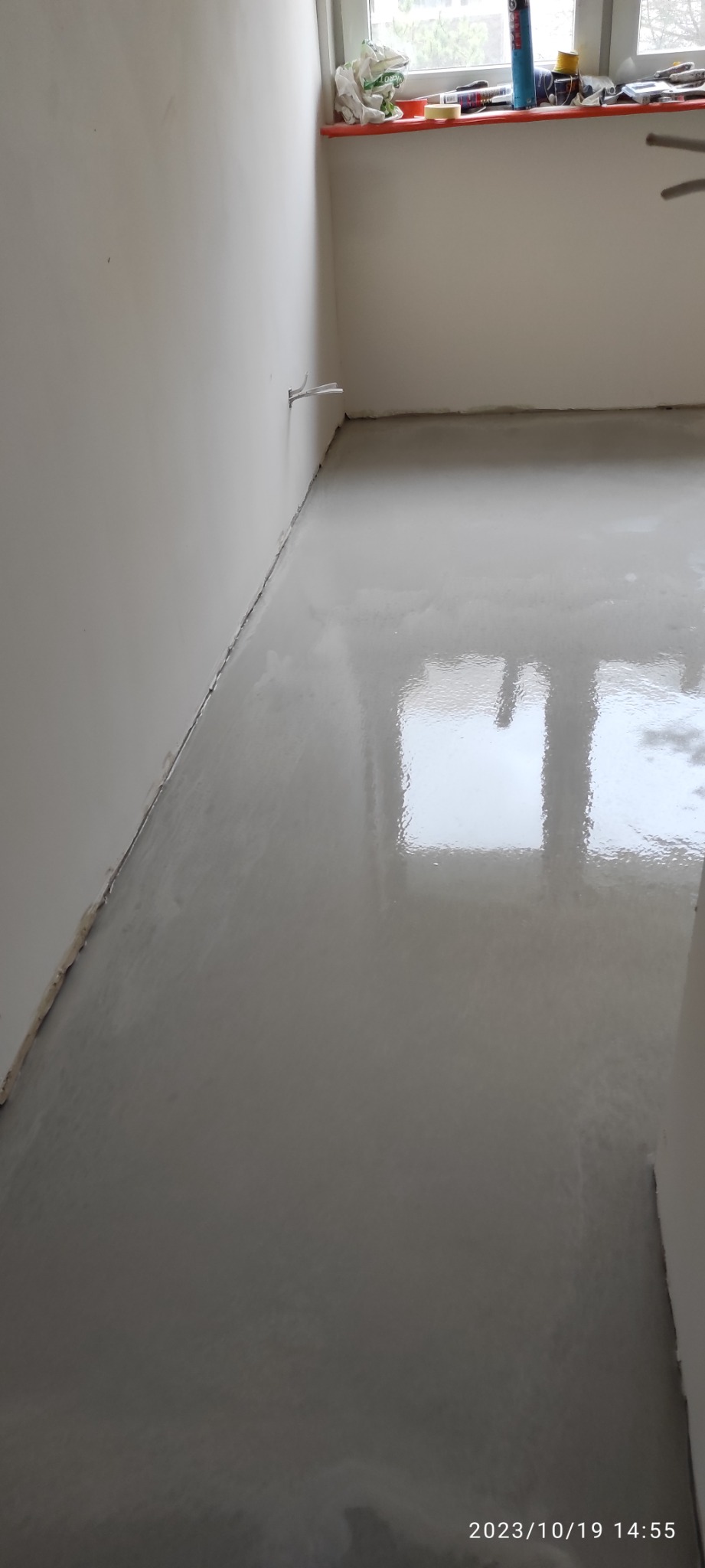 Wnętrze pokoju z wylewką samopoziomującą na podłodze, widoczne kable wystające ze ściany, okno z narzędziami na parapecie.