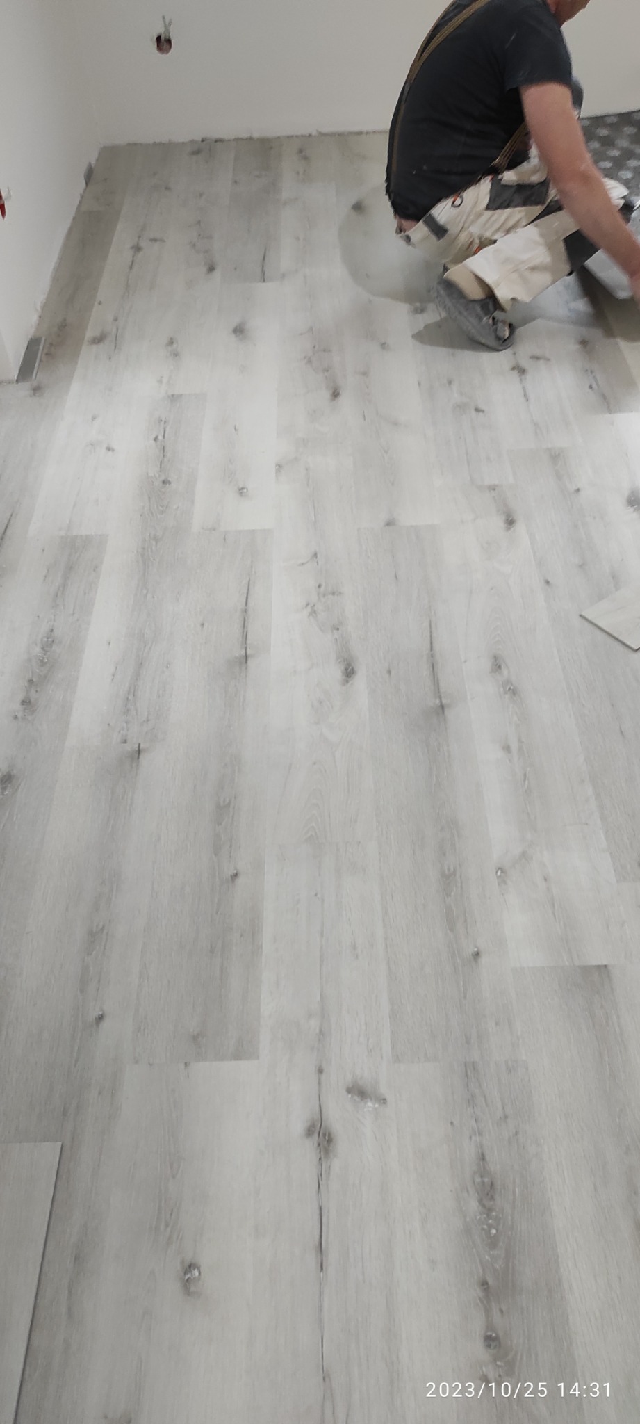 Pracownik w kombinezonie montuje jasnoszare panele podłogowe z widoczną strukturą drewna, na częściowo wyłożonej podłodze w jasnym pomieszczeniu.