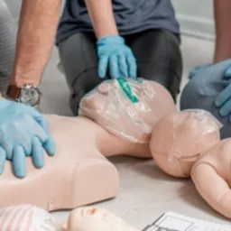 Instruktorzy w niebieskich rękawiczkach demonstrują resuscytację krążeniowo-oddechową na manekinach niemowląt i dorosłych podczas szkolenia.