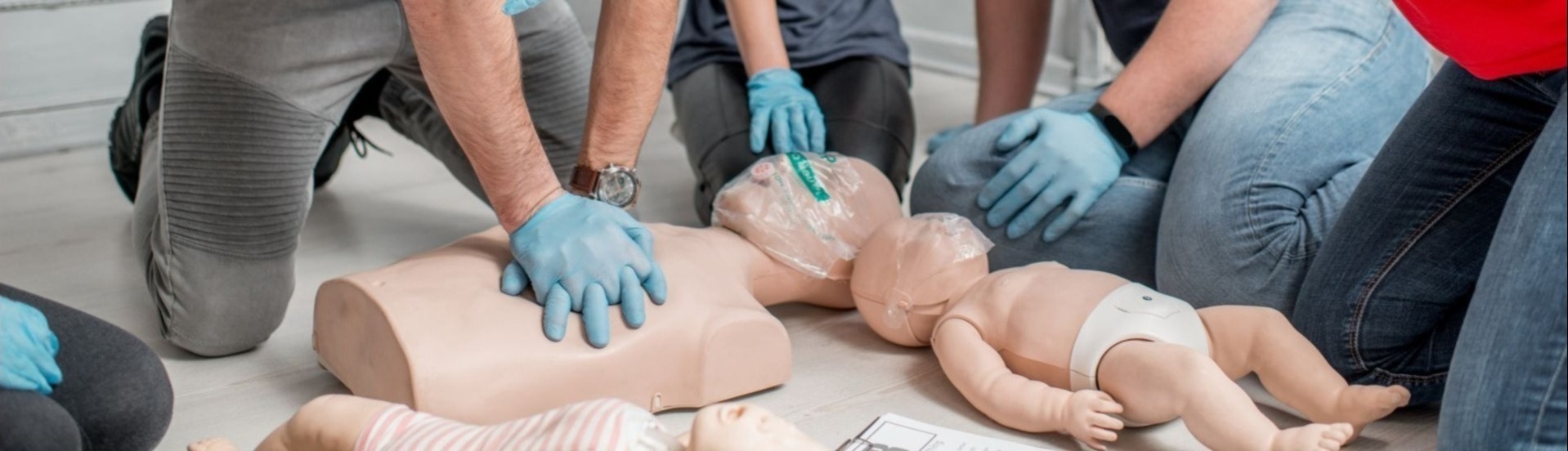 Instruktorzy w niebieskich rękawiczkach demonstrują resuscytację krążeniowo-oddechową na manekinach niemowląt i dorosłych podczas szkolenia.