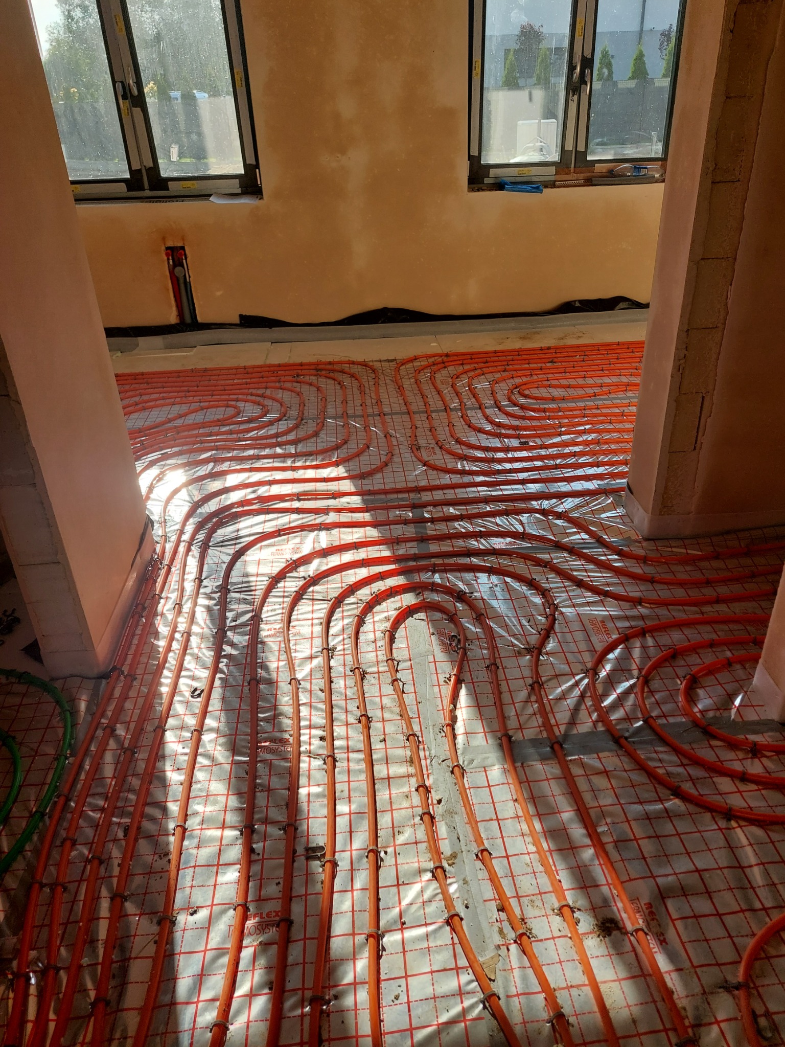 Pomieszczenie w trakcie montażu ogrzewania podłogowego, widoczne rury grzewcze w kolorze pomarańczowym ułożone na folii izolacyjnej z siatką, fragmenty ścian w stanie surowym, okna z widokiem...