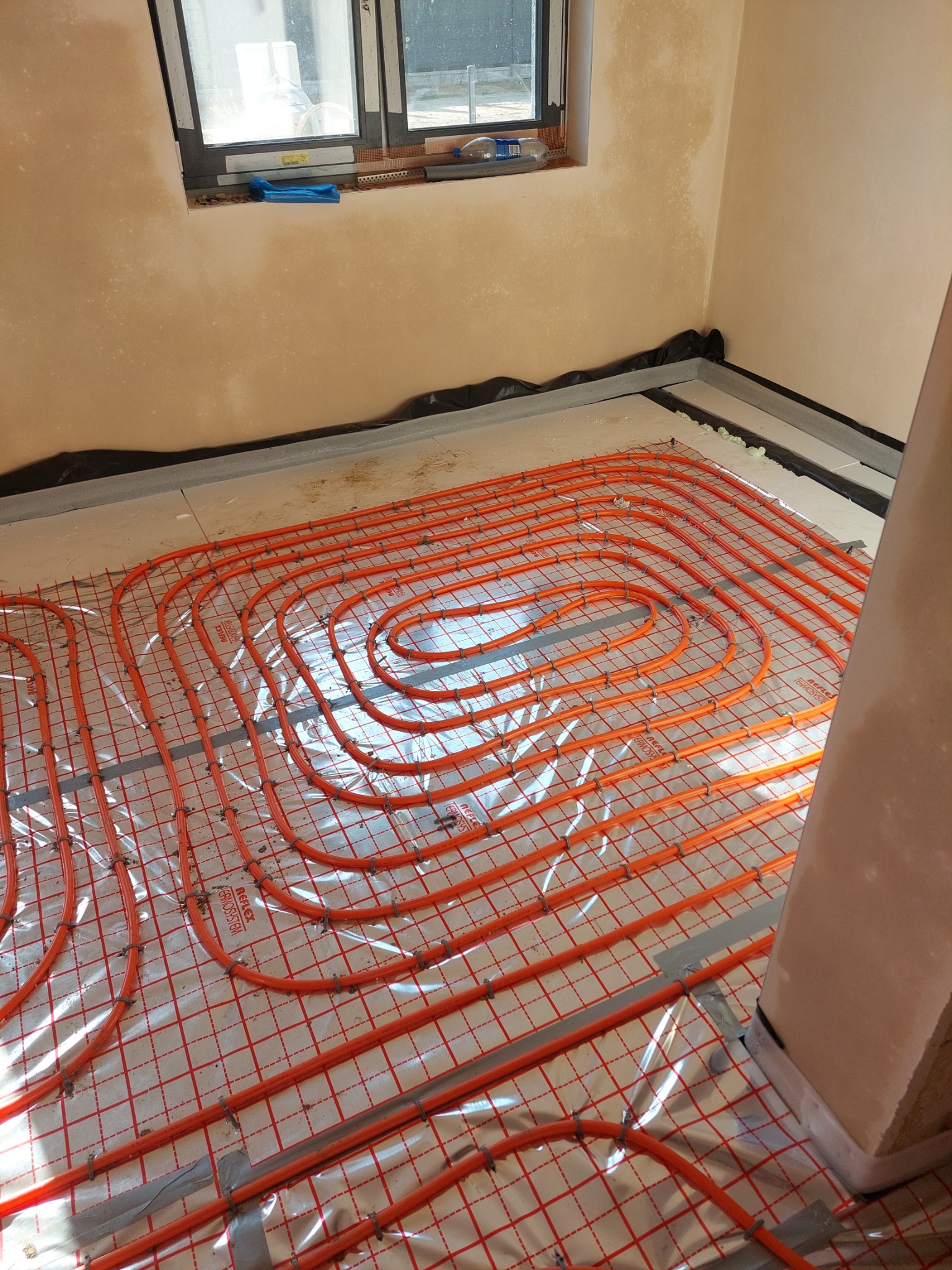 Pomarańczowe rury ogrzewania podłogowego ułożone spiralnie na folii z czerwoną siatką w surowym wnętrzu z oknem i przygotowanymi ścianami.