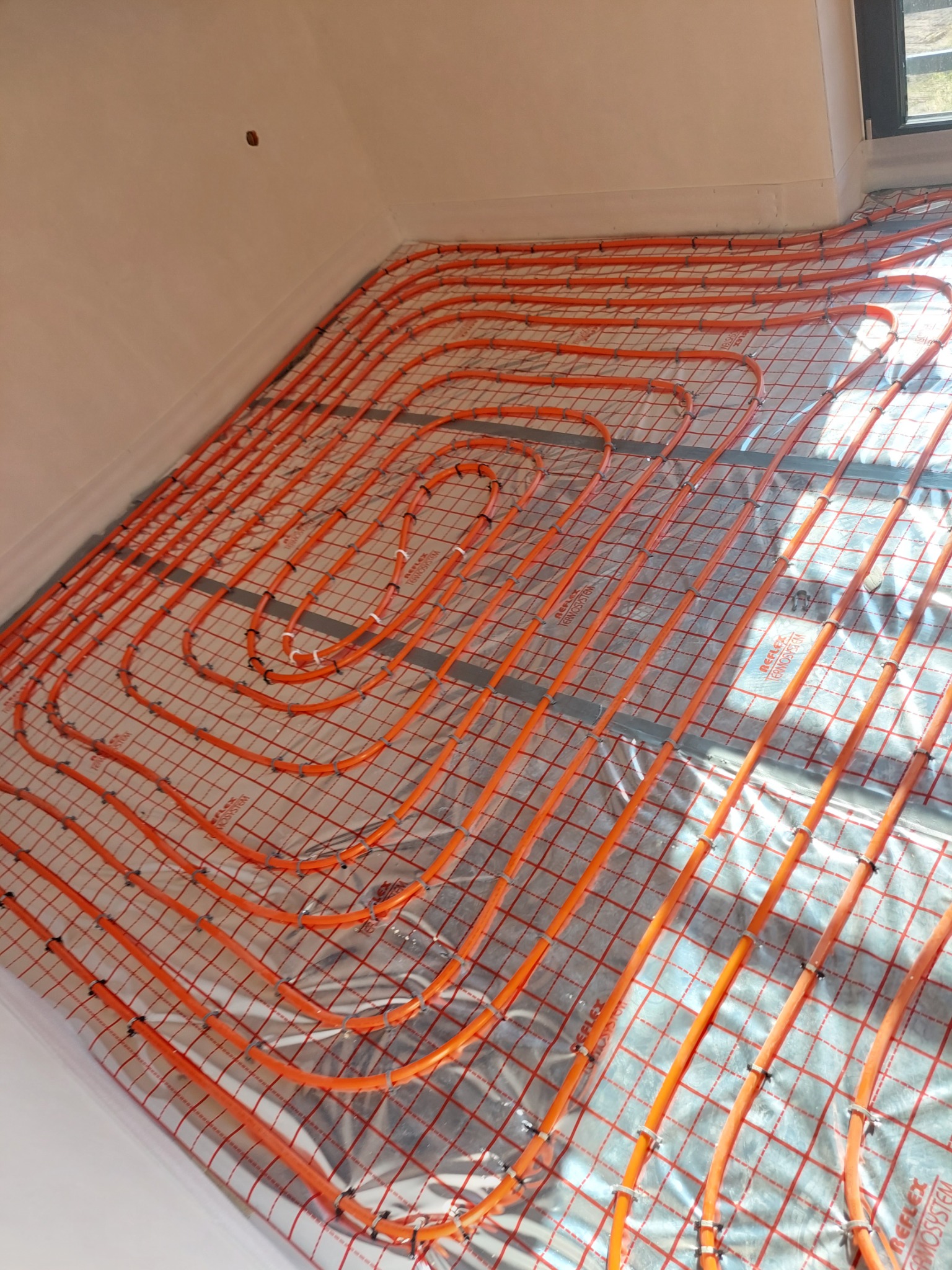 Pomarańczowe rury ogrzewania podłogowego ułożone w pętle na srebrnej folii z czerwoną siatką w nowym budynku, widoczne białe ściany i okno.