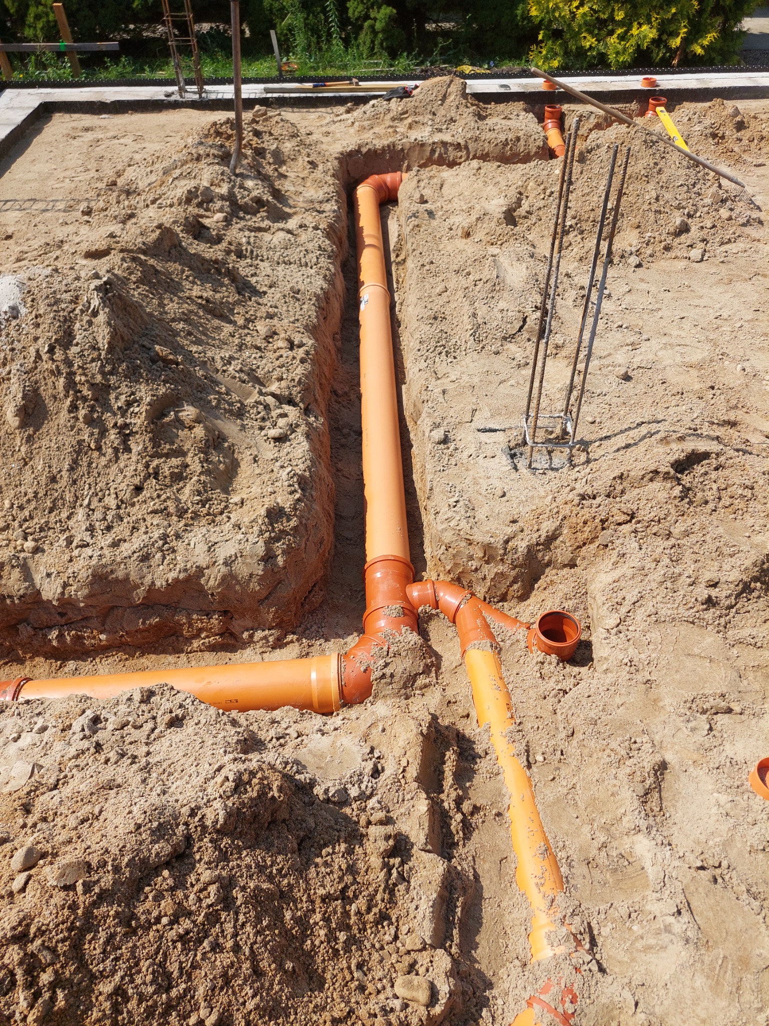 Pomarańczowe rury kanalizacyjne ułożone w wykopie w ziemi, widoczne złączki i fragmenty rur, elementy konstrukcyjne w tle.