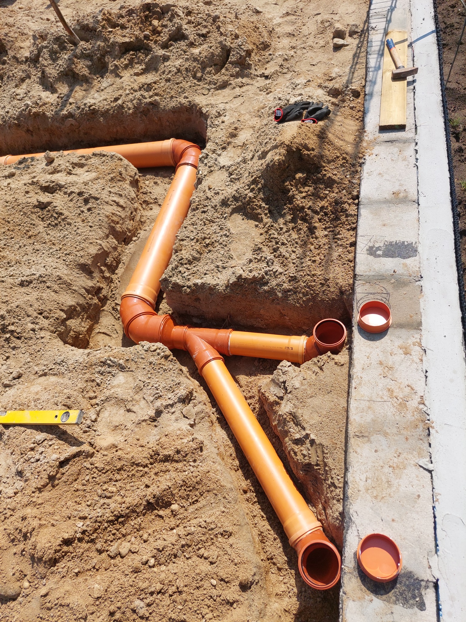 Pomarańczowe rury kanalizacyjne ułożone w wykopie ziemnym, widoczna poziomica, rękawice robocze i młotek na desce przy betonowym obrzeżu.