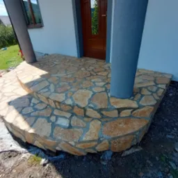 Dwu-stopniowe schody wejściowe do domu wyłożone nieregularnymi kamieniami w odcieniach beżu i brązu, z szarymi filarami po bokach i brązowymi drzwiami wejściowymi w tle.